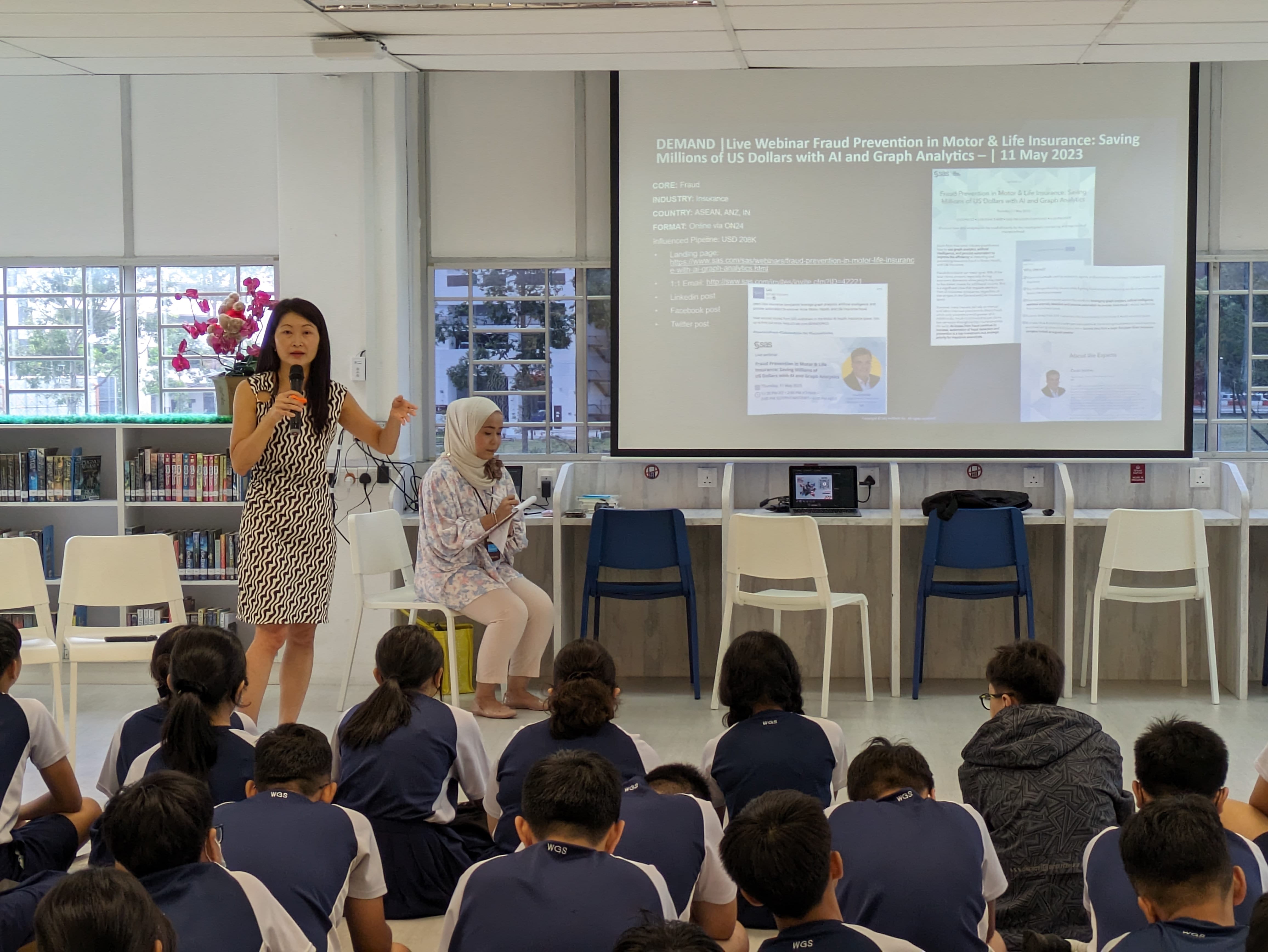A woman speaks with a microphone to students seated on the floor, with a projected slide about fraud prevention visible behind her.